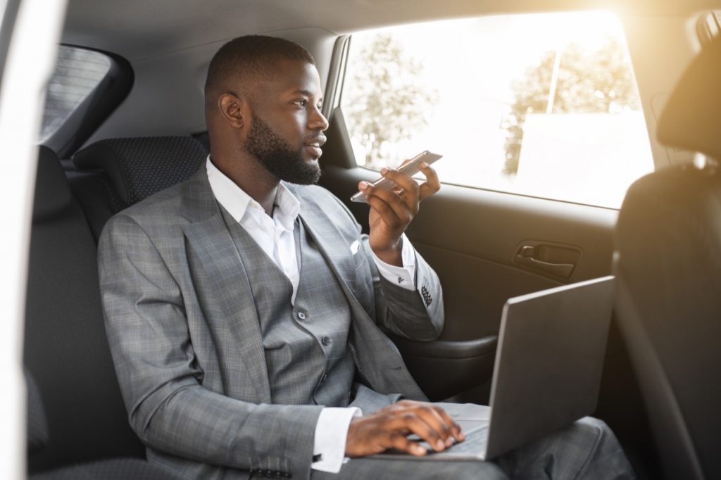 Young black manager working on laptop while going to office by luxury car, recording audio message for his assistant on mobile phone, sitting on car back seat, copy space, sun flare By Prostock-studio