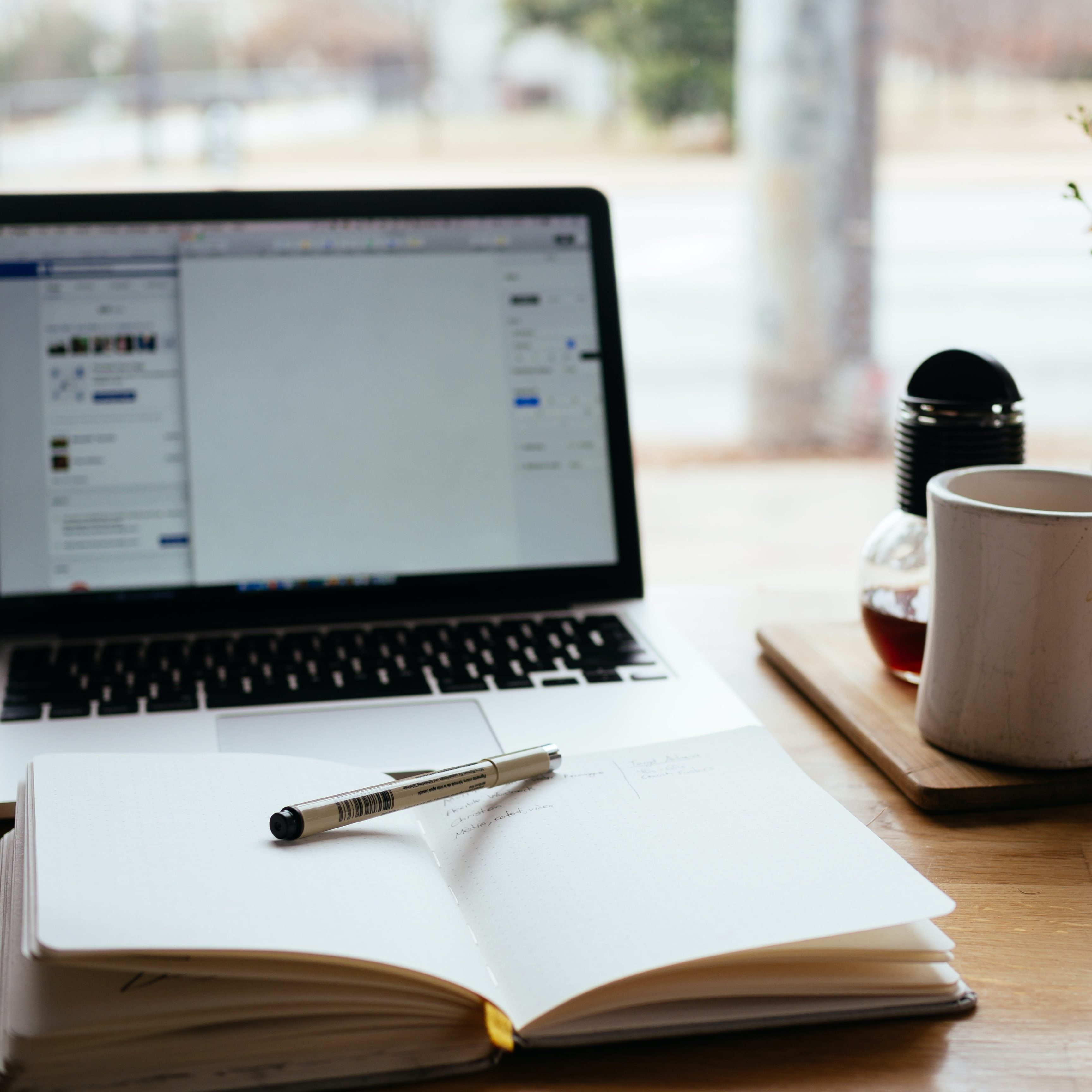 A note book with a pen on top with a laptop open behind to Facebook. A tea mug to the right of the computer. A blurry outdoor background.