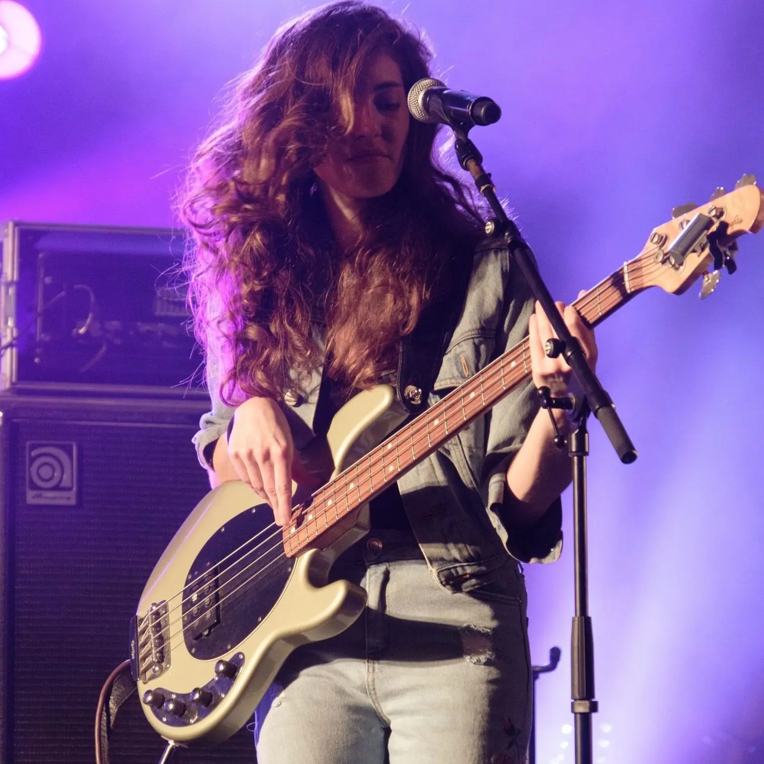 Woman playing a live performance on stage. Standing behind a microphone playing a bass guitar. Her brown hair is covering most of her face. Bright purple lights shine behind her.