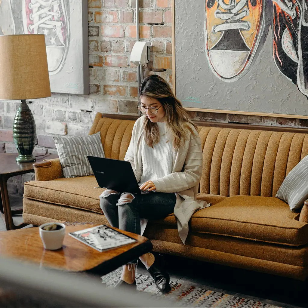 Asian woman sitting on brown couch in a funky office space with a lamp to her right, a wooden table in front of her and an artwork of shoes behind. She is on a black laptop wearing a white top with a grey cardigan and black pants. She has brown hair with blonde tips and glasses.