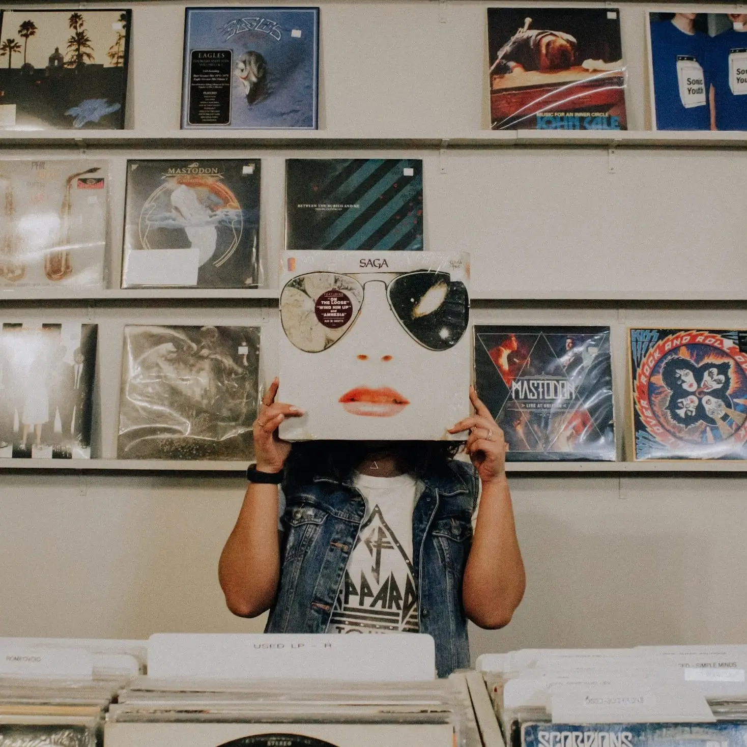 woman holding album cover artwork in front of her face. The artwork has sunglasses a nose and a mouth where hers should be. She is wearin a denim shirt and a white t-shirt with black design on it. there is a wall of vinyl behind her.