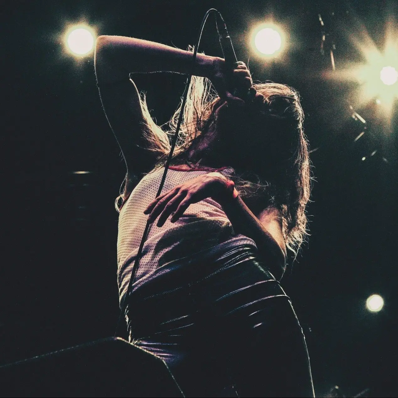 Female vocalist doing a live performance. She is holding a microphone above her face. Wearing a white singlet and black pants. The background is black and dark except for some yellow lights that are twinkling in the background.