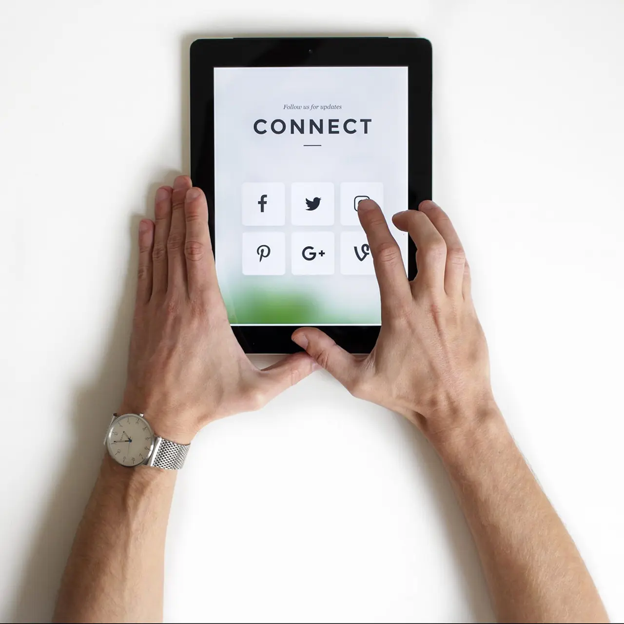 Two hands holding an iPad. The iPad says connect at the top of the screen and has icons to login online to Facebook, Twitter, Intagram, Pinterest, Google and Vine. One of the fingers is reaching to tap on Instagram.