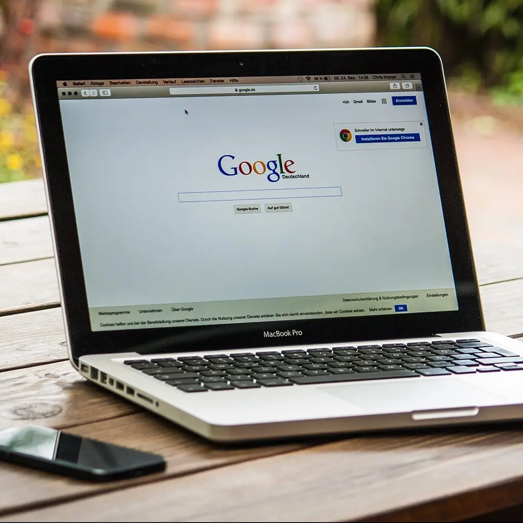 Google website open on mac laptop on top of a wooden table with a phone next to it.