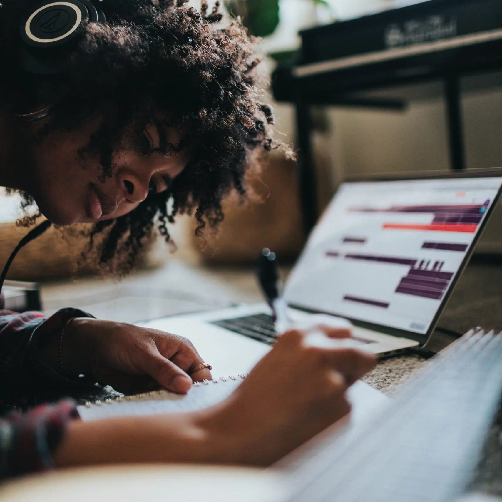 Black woman with headphones on writing something on a piece of paper. Wild curly brown hair. Guitar and laptop open on a music program.