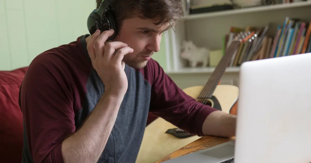 Online music distribution setup with a man using headphones and a laptop, with a guitar in the background.
