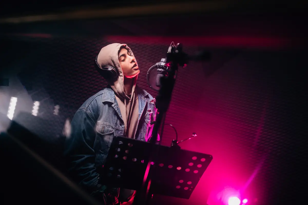 Live streaming for musicians with a hoodie-wearing singer recording vocals under colorful studio lights