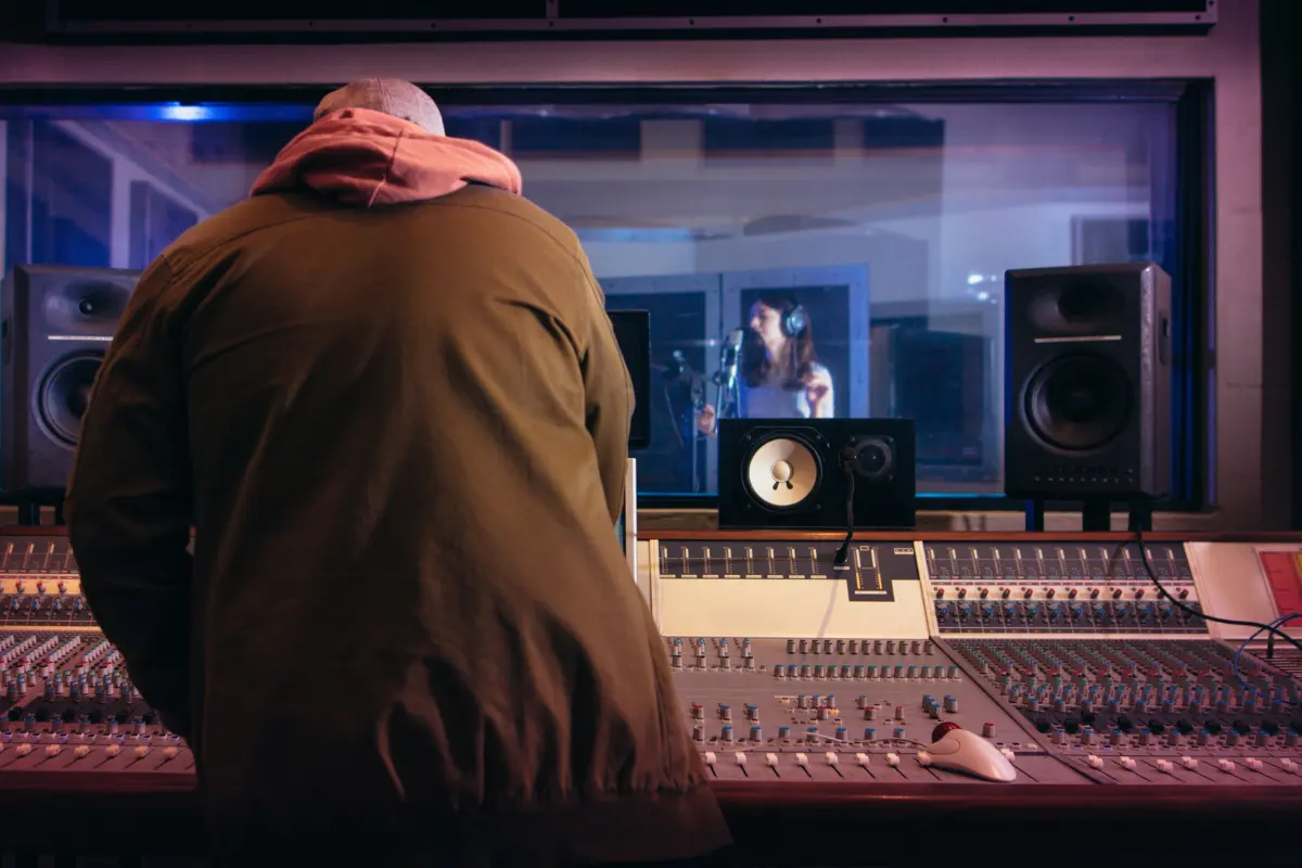 Music marketing online with a producer monitoring a recording session through studio glass during a vocal performance.