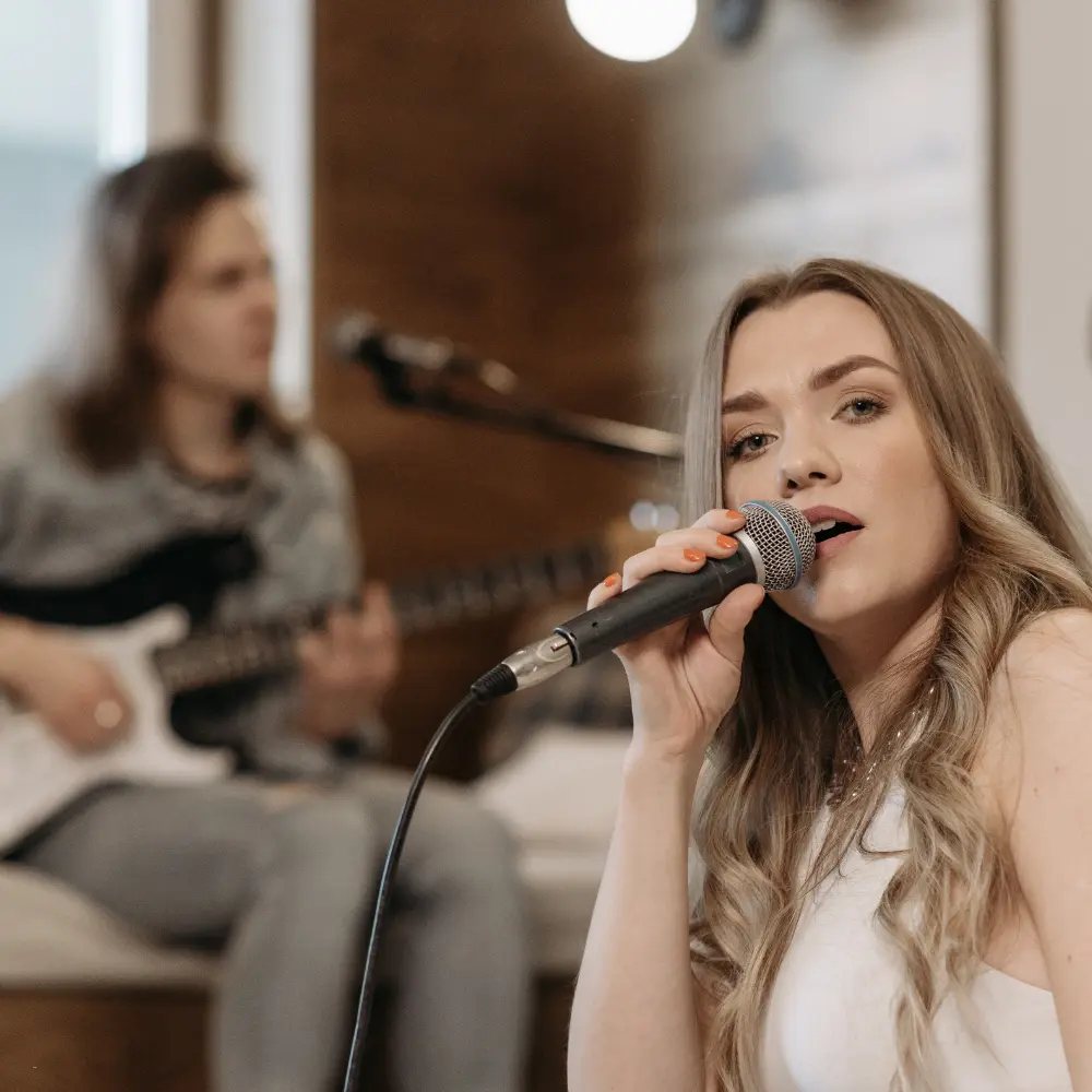 Remote music collaboration shown with a vocalist singing while a guitarist plays in a cozy indoor setting