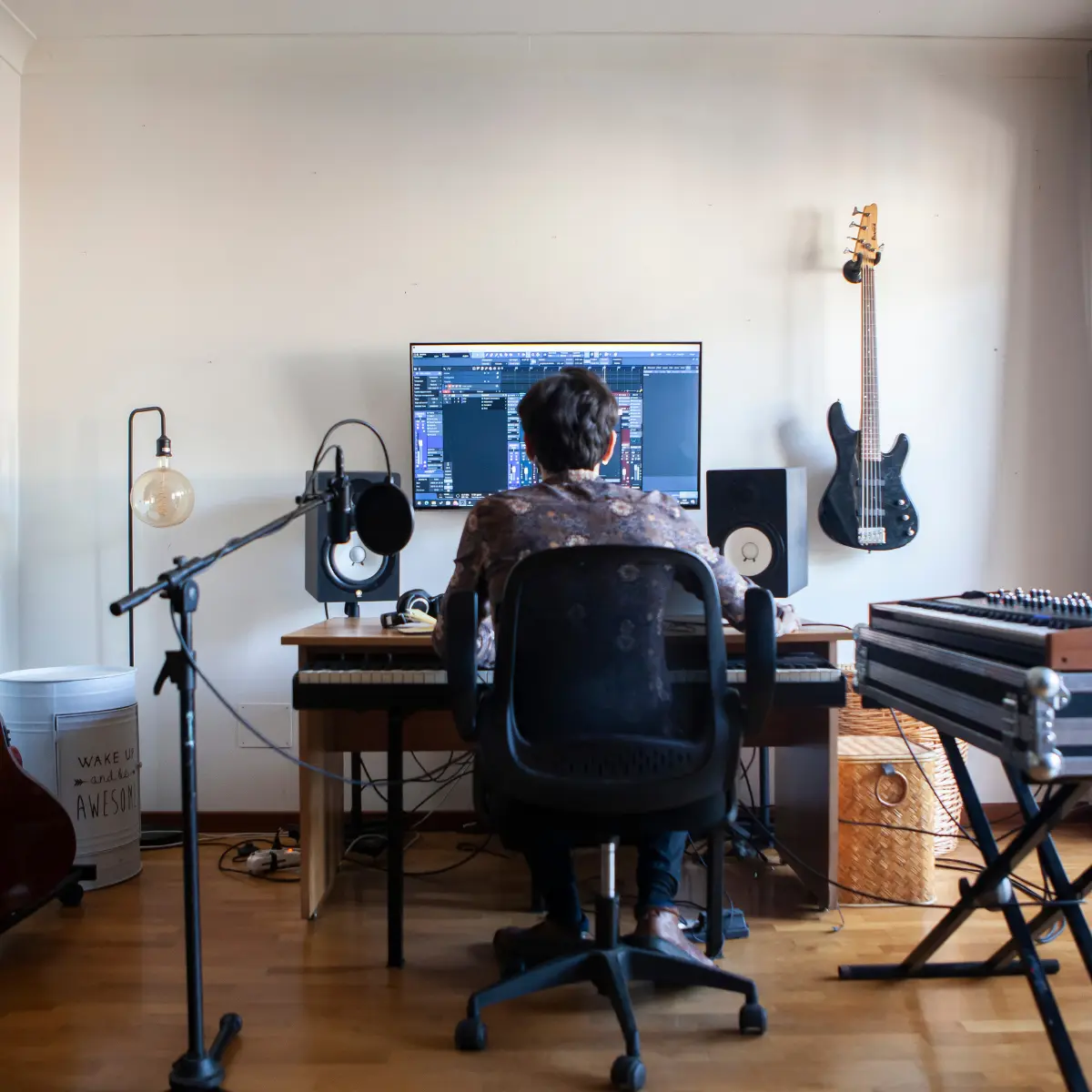 Music producer working at a home studio setup with a digital audio workstation and bass guitar on wall.